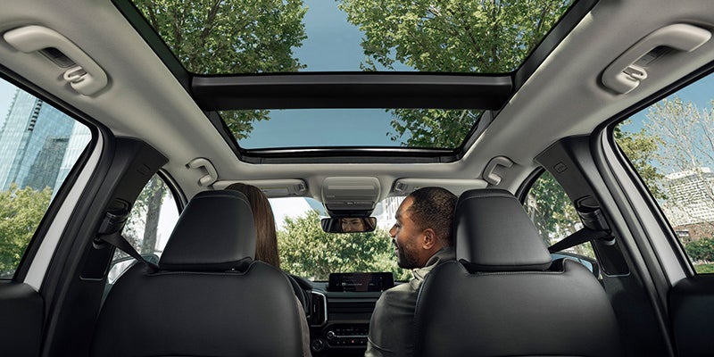 Interior of a car looking forward, showing panoramic sunroof, two people in front seats, and trees outside.