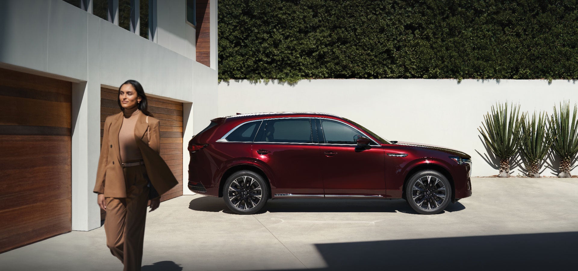 A confident woman in a tan suit walks past a glossy red SUV parked in a modern driveway.