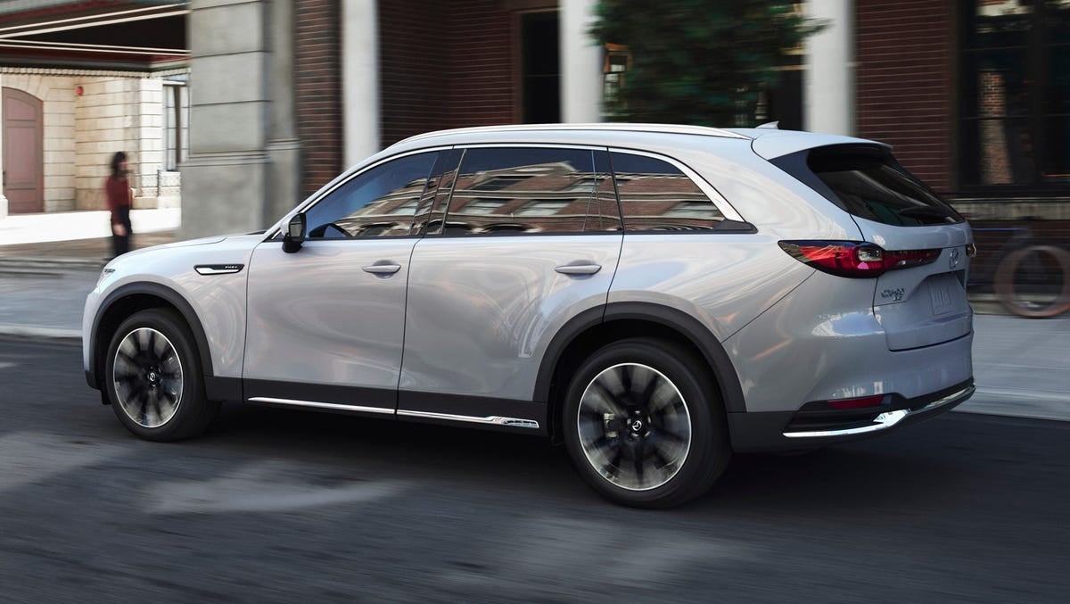 A light gray Mazda CX-90 SUV driving down a city street with blurred surroundings.