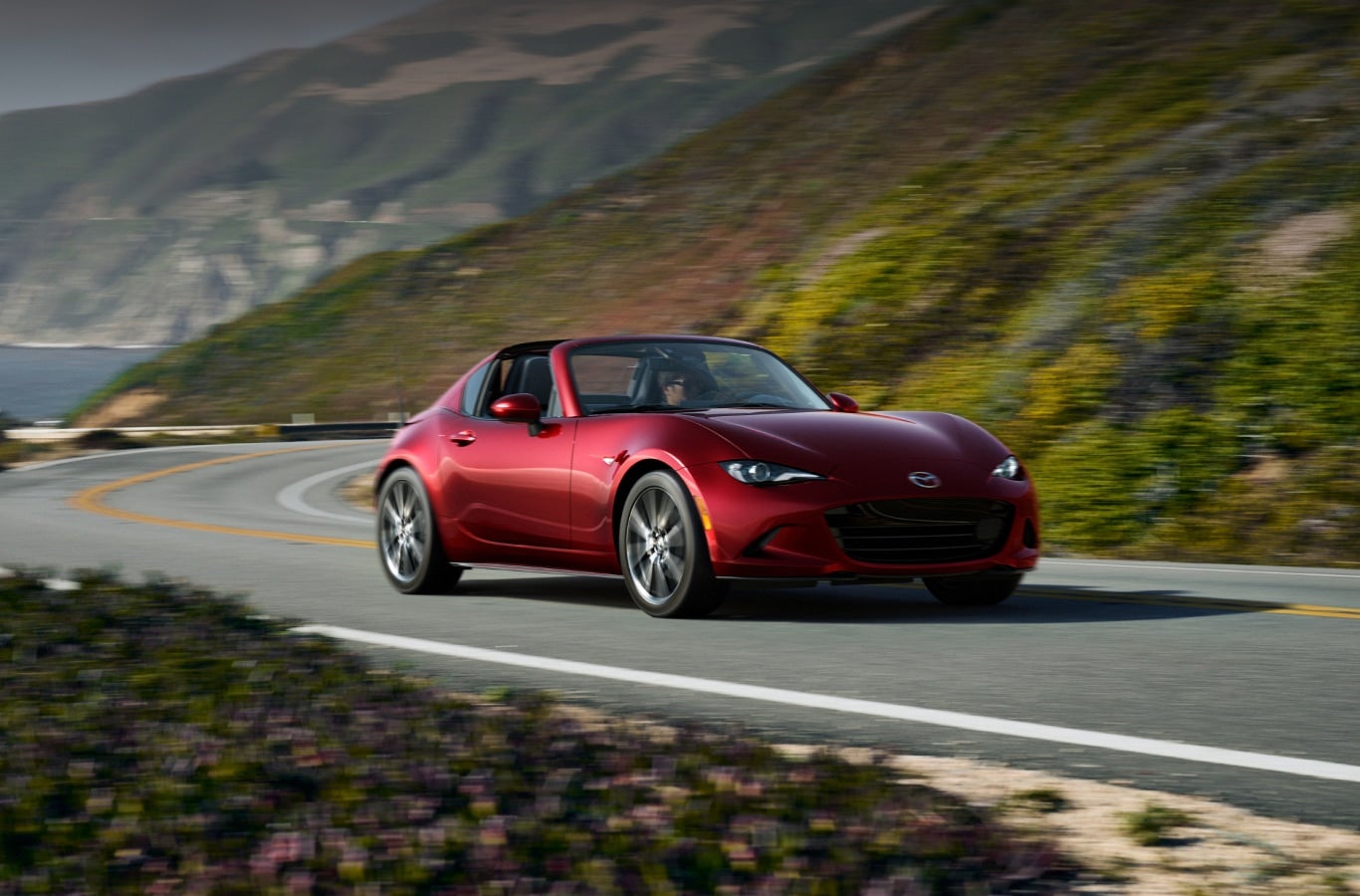 A red Mazda MX-5 Miata RF hardtop convertible taking a sharp turn on a scenic coastal highway.
