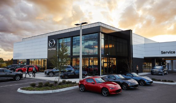 A modern Mazda car dealership building with large glass windows and parked cars out front under a cloudy sunset sky.