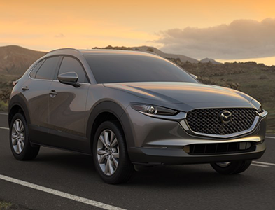Gray Mazda CX-30 SUV driving on a road at sunset with mountains in the background.