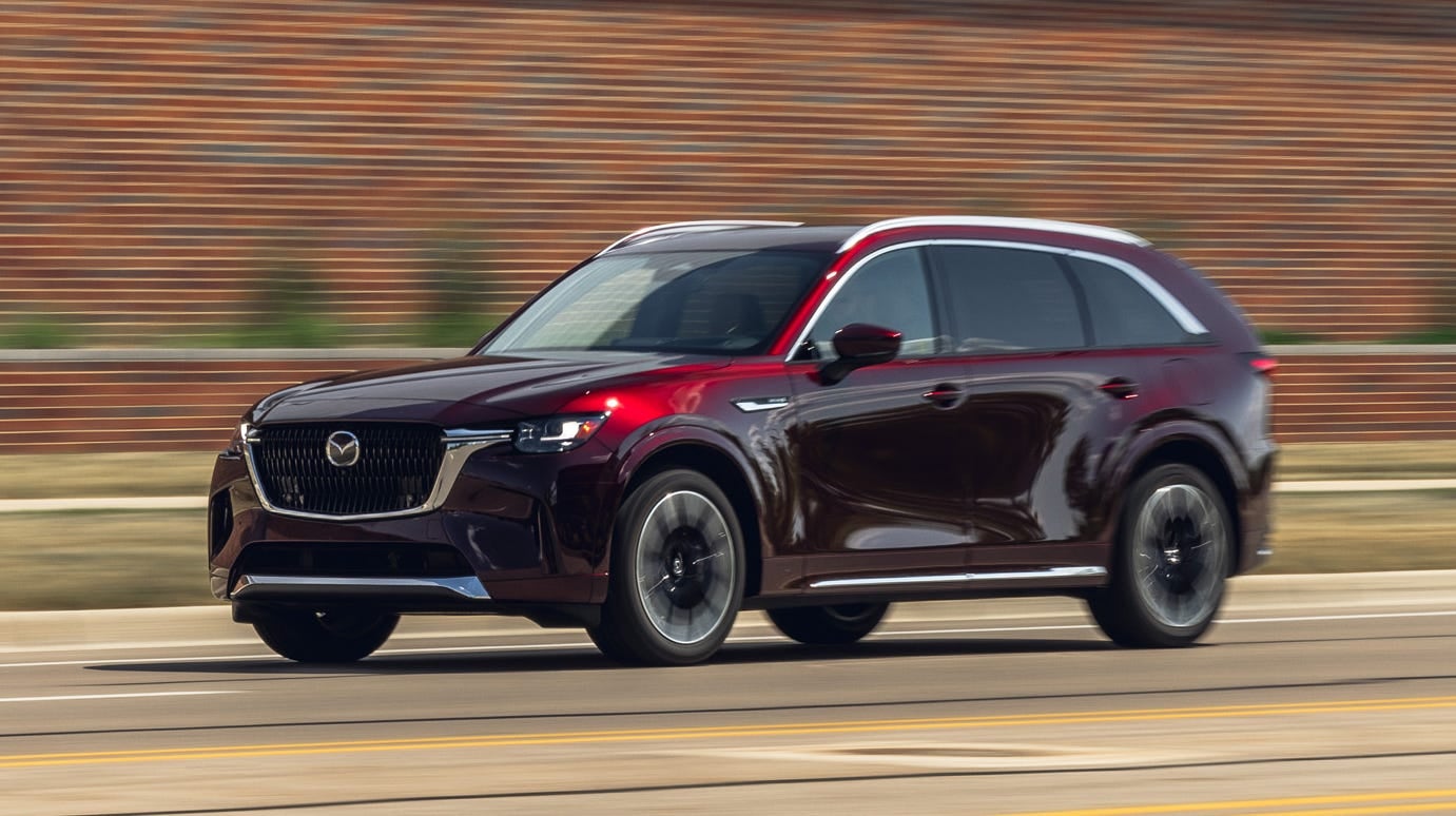 A dark cherry red Mazda CX-90 SUV drives left on a road, with a blurry brick building in the background.